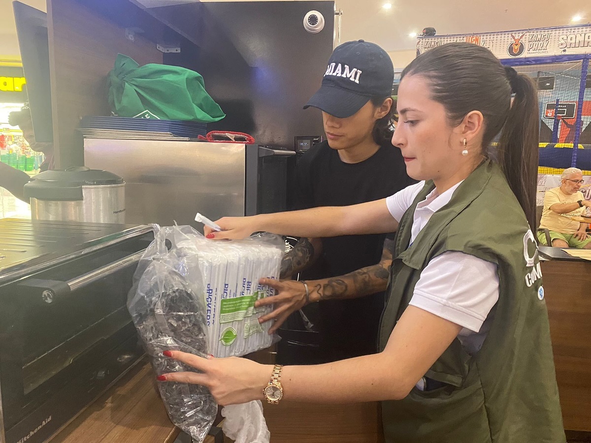 Dos personas inspeccionan y organizan paquetes de materiales biodegradables en un local de alimentos. Una lleva un chaleco oficial y parece estar supervisando el procedimiento.