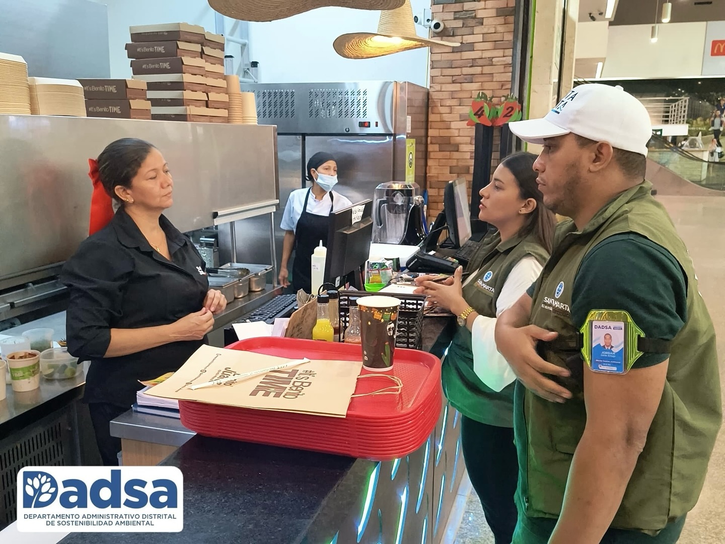 Inspectores dialogan con una empleada en un local de comida, destacando la importancia de prácticas sostenibles en el uso de empaques y utensilios.