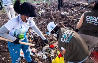 Dos personas recogiendo basura en un área de residuos, usando herramientas de limpieza y guantes protectores.