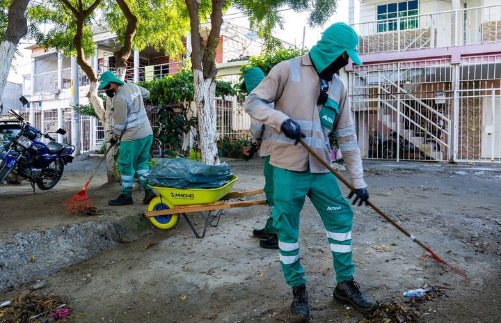 Trabajadores de limpieza barrriendo las calles, con uniformes verdes y herramientas de trabajo como carretillas y escobas.