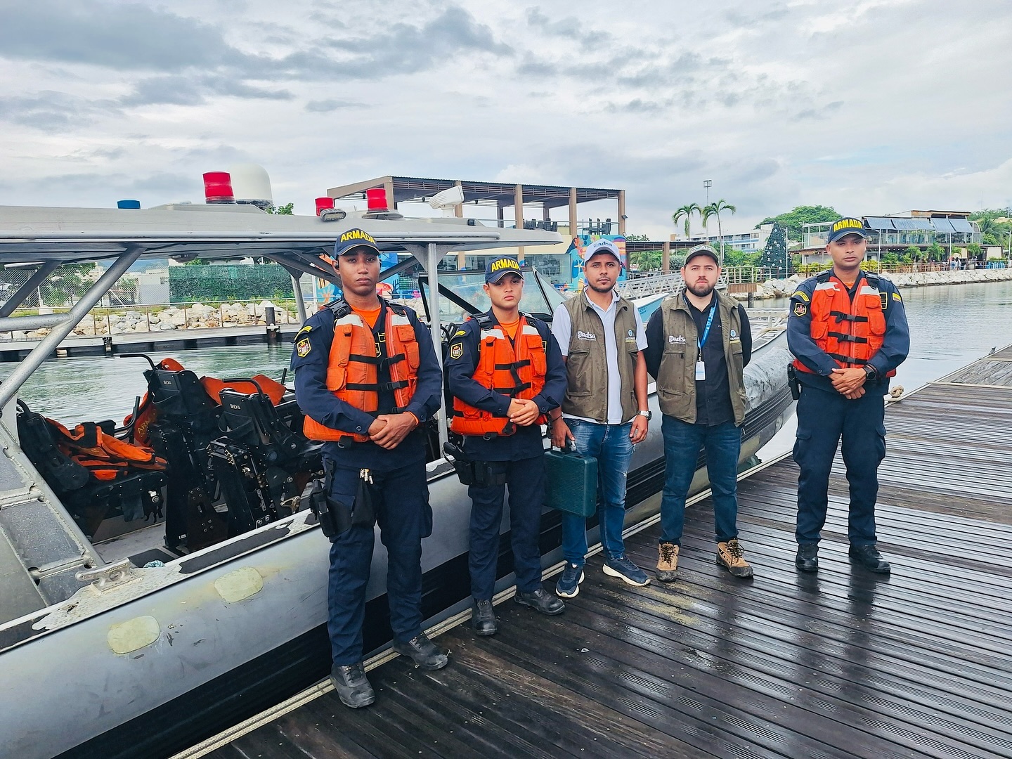 Grupo de cinco personas posando junto a una embarcación en un muelle. Tres de ellos pertenecen a la Armada, vistiendo uniformes azules con chalecos salvavidas naranjas, mientras que dos llevan chalecos distintivos de una entidad administrativa. Al fondo, se observan construcciones y áreas verdes en un ambiente costero.