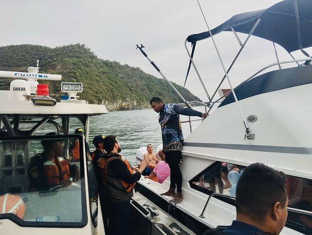 Personas a bordo de una lancha participan en una operación de rescate en el mar, mientras un hombre es asistido por miembros de seguridad marina con un paisaje montañoso al fondo.